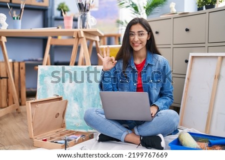 Young teenager girl sitting at art studio using laptop doing ok sign with fingers, smiling friendly gesturing excellent symbol 
