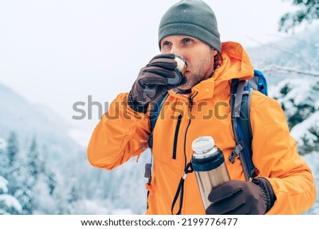 Man drinking a hot drink from thermos flask dressed bright orange softshell jacket while he trekking winter mountains route. Active people in the nature concept image. Royalty-Free Stock Photo #2199776477