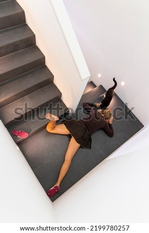 Attractive blonde woman in dark dress fell down the stairs and the black cat passed her. The pink slippers have flown. Copy space
