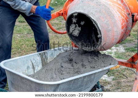 A worker pours cement mortar, concrete, from a concrete mixer into a wheelbarrow. Preparation of cement mortar at the construction site Royalty-Free Stock Photo #2199800179
