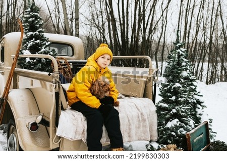 cute teenage boy in winter coat and knitted hat and pet poodle dog sit near beige retro pickup truck decorated for Christmas and New Year vintage interior suitcases, skis, sledge