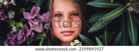 portrait face of candid beautiful little kid girl of eight years old with brown eyes on background of green plants and pink flowers during a summer vacation travel. gen z mental health concept. banner