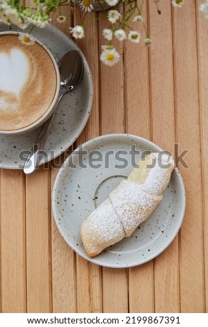 French croissant roll under powder. Cappuccino in white ceramic cup and blurred foreground of chamomile flowers on wooden table at the terrace. Aesthetic breakfast.