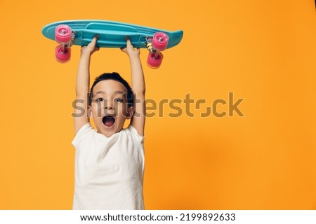 a little boy of preschool age is standing on an orange background in a white T-shirt, smiling fervently holding a skate above him with his mouth open in surprise and looking away
