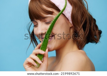 a sweet, happy woman stands on a blue background holding a sprig of aloe vera in her hand and presenting it to her face, smiling happily, closing her eyes with pleasure