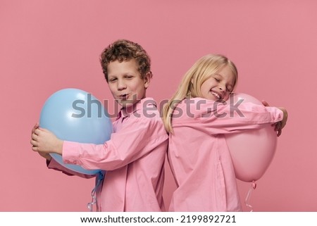 cute, happy kids in pink clothes on a pink background stand sideways to the camera and hug their big pink and blue balloons. Horizontal studio photo with empty space for advertising text insertion