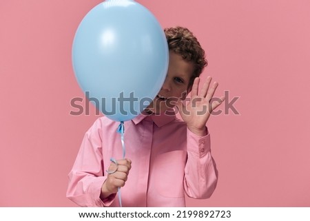a cute, funny boy of school age stands on a pink background in pink clothes and funny hides his face behind a big blue balloon and waves his hand happily smiling at the camera.