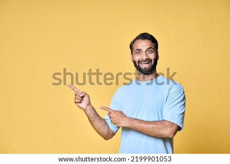 Happy excited indian man looking at camera pointing aside with fingers hand gesture indicating advertising new product commercial promo, presenting sale offer standing isolated on yellow background. Royalty-Free Stock Photo #2199901053