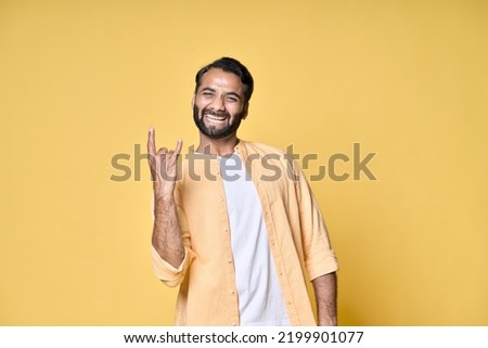 Happy funky cool indian man rocker punk hipster showing rock n roll heavy metal horns hand gesture screaming having fun advertising party festival isolated on yellow background.