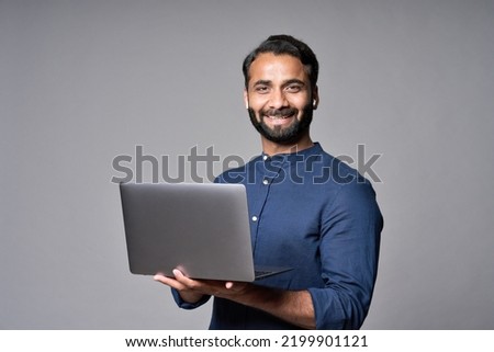 Happy smiling indian business man employee or manager standing isolated on gray background holding laptop advertising online products, business trainings and webinars, websites or services. Copy space Royalty-Free Stock Photo #2199901121