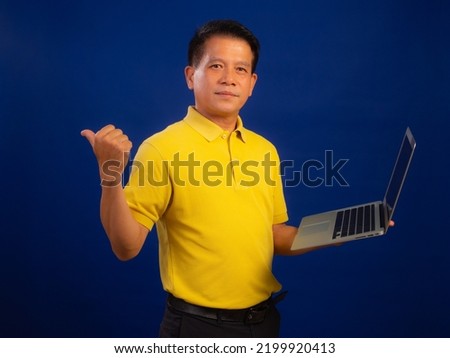 Portrait of middle-aged asian man in casual yellow T-shirt holding laptop isolated on blue background in studio, looking at camera