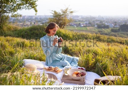 Portrait of woman in a long dress with short hair sitting on a white blanket with fruits and pastries and looking at the pictures on camera. Concept of having picnic during summer holidays or weekends