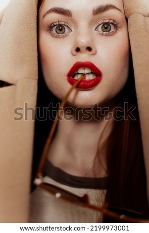 A playful beautiful girl with a coat on her head bites the bow of her glasses. Close-up portrait