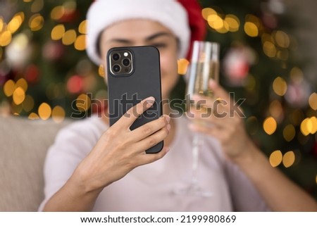 Happy female smartphone user taking selfie with New Year flute of wine at glowing tree in background, making video call for Christmas congratulation, giving wish. Mobile phone in hand close up