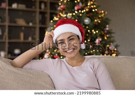 Happy joyful Indian girl in Santa hat looking at camera, smiling, relaxing on couch with blurred glowing Christmas tree lights in background, celebrating New Year. Head shot portrait