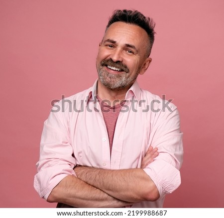 Smiling man posing with arms crossed on pink background. Portrait of attractive handsome mature man in rose shirt with crossed arms. Healthy happy smile senior beard caucasian man isolated on pink.
