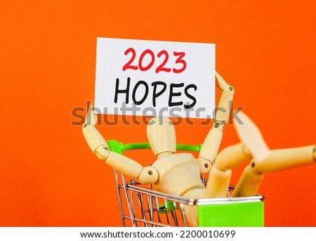 White paper with words 2023 Hopes, human model in shopcart. Beautiful orange table orange background. Business and 2023 hopes concept. Copy space.