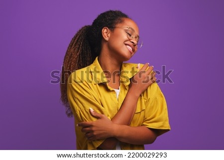 Young attractive African American woman hugging herself closing eyes in pleasure and smiling feeling happy dressed in casual clothes stands on purple studio background. Selfishness, loneliness concept Royalty-Free Stock Photo #2200029293