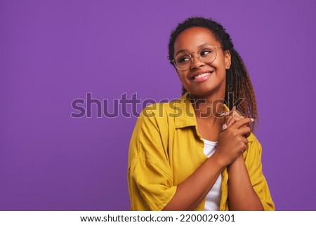 Young cheerful African American woman millennial look around and smile when see friend or great promotional offer dressed in casual oversize shirt stands on purple studio background Royalty-Free Stock Photo #2200029301