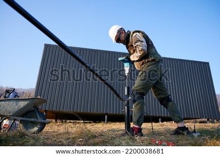Male worker building pile foundation for wooden frame house. Man builder in white safety helmet drilling piles into the ground on blue sky background.