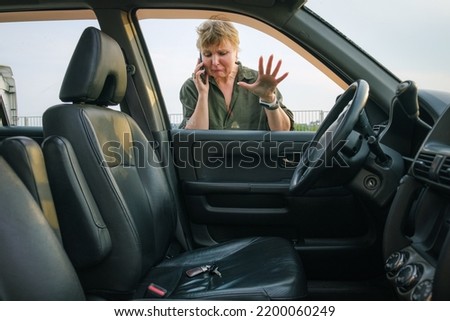 Middle-aged woman looks through the glass into the interior of her car with the keys in the driver's seat. Woman driver forgot her keys in the car and calling technical assistance Royalty-Free Stock Photo #2200060249