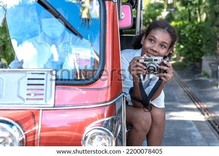 Young independent tourist girl sitting beside Tuk Tuk taxi driver holding camera taking pictures, tuk tuk travelling during summer vacation in Chiang Mai, Thailand