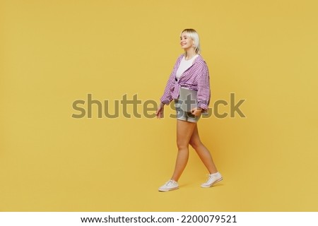 Full body side view young programmer student blonde IT woman 20s wear pink tied shirt white t-shirt hold closed laptop pc computer walk go isolated on plain yellow background. People lifestyle concept