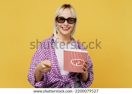 Young blonde smiling cheerful happy satisfied woman in 3d glasses watch movie film hold bucket of popcorn in cinema point index finger camera on you isolated on plain yellow background studio portrait