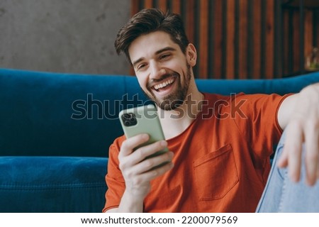 Close up young man wears red t-shirt hold in hand use mobile cell phone sit on blue sofa stay at home hotel flat rest relax spend free spare time in living room indoors grey wall People lounge concept