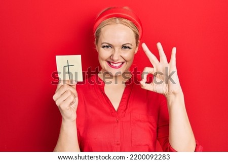 Young blonde woman holding catholic cross reminder doing ok sign with fingers, smiling friendly gesturing excellent symbol 