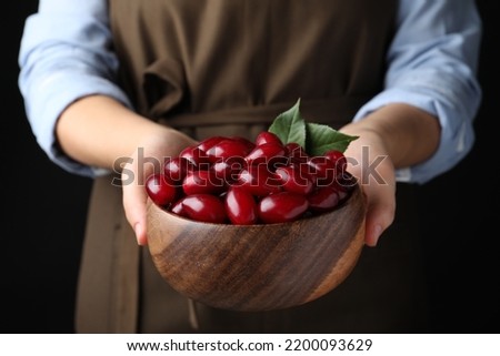 Woman with wooden bowl of fresh ripe dogwood berries on black background, closeup