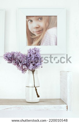 Small bouquet of lilac in glass bottle on white wooden shelf with photo frame on it as detail of interior
