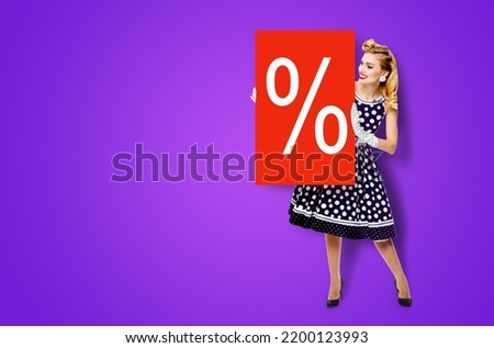 Beautiful woman in pinup dress holding, showing red board with procents % sign. Full body pin up girl with advertising offer, isolated on violet purple background. Sales, discounts rebates ad concept.