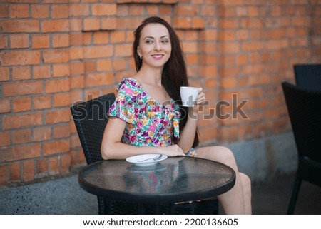 Stylish slender smiling woman with long dark hair, with a cup of coffee, in a light summer dress with a floral pattern, sits at a table in a cafe on the street, against the backdrop of a brick wall.