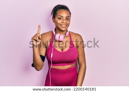 Young african american girl wearing gym clothes and using headphones pointing finger up with successful idea. exited and happy. number one. 