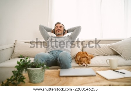 Calm Middle age Caucasian man sitting on sofa listening to music enjoying meditation for sleep and peaceful mind in wireless headphones, leaning back with his lovely chihuahua dog sit besides. Royalty-Free Stock Photo #2200146255
