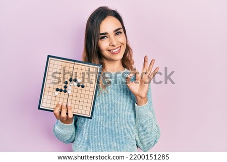 Young hispanic girl holding asian go game board doing ok sign with fingers, smiling friendly gesturing excellent symbol 