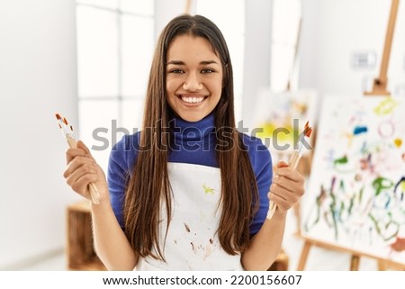Young latin woman smiling confident holding paintbrushes at art studio