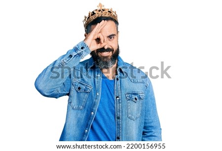 Hispanic man with beard wearing king crown smiling happy doing ok sign with hand on eye looking through fingers 