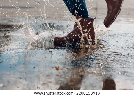 Woman wearing rain rubber boots walking running and jumping into puddle with water splash and drops in autumn rain. Royalty-Free Stock Photo #2200158913