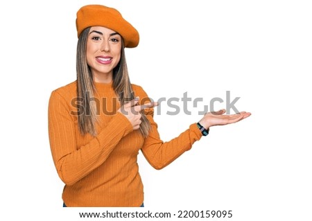 Young hispanic woman wearing french look with beret amazed and smiling to the camera while presenting with hand and pointing with finger. 