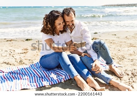 Middle age hispanic couple make selfie by the smartphone sitting on the towel at the beach.