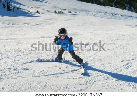 a little beginner skier on a ski slope in a funny inverted position and smiling behind the goggles looking at the camera, child having fun learning to ski, horizontal Royalty-Free Stock Photo #2200176367