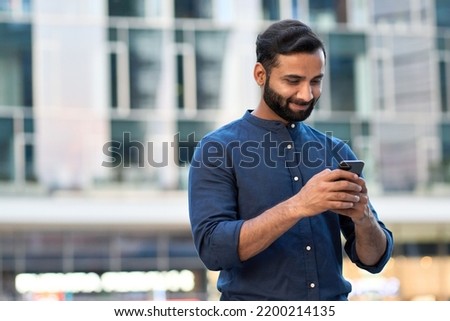 Smiling indian business man, eastern businessman holding cell phone using smartphone mobile apps texting message, surfing social media tech standing in urban city on modern street outdoors. Royalty-Free Stock Photo #2200214135