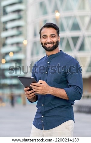 Smiling indian business man professional executive manager holding digital tablet online fintech outside, happy eastern businessman ceo using pad standing outdoors in big urban city street, vertical. Royalty-Free Stock Photo #2200214139
