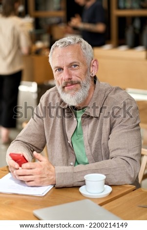 Middle aged gray-haired older adult business man, mature bearded male professional businessman holding smartphone using cell phone working, mobile learning sitting at cafe table. Vertical portrait