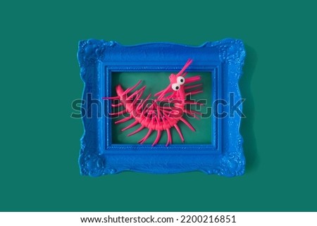 A pink plastic caterpillar with big eyes in a retro blue frame against green background. Funny surreal concept for Halloween celebration party invitation or advertisement