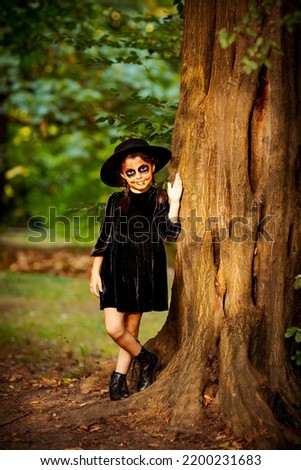 a small, beautiful girl in a black dress and hat, a face in Halloween makeup, stands near a tree in the park