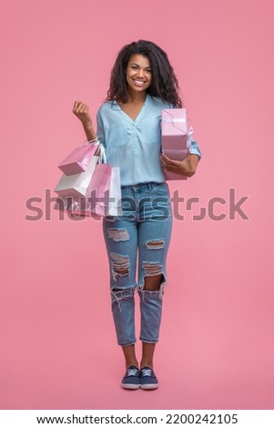 Studio shot, full-length vertical portrait of happy smiling beautiful casually dressed young woman posing standing with gift box and bunch of shopping bags in hands over pastel pink background.