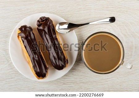 Two eclairs with chocolate glaze in white glass plate, teaspoon, transparent cup of coffee with milk on wooden table. Top view
